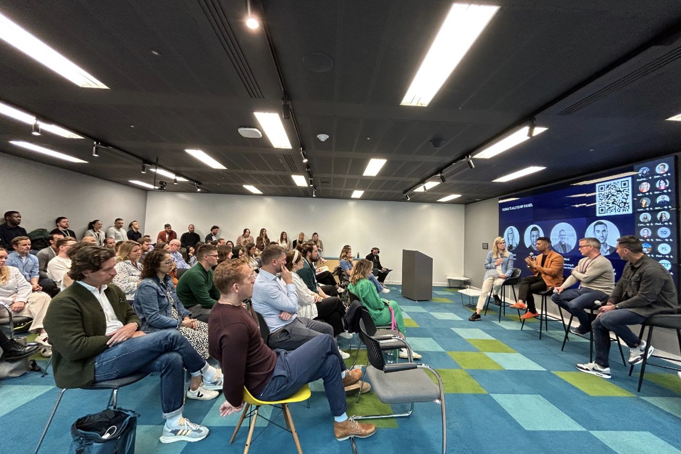 A panel of four seated at the front of a full room, the audience in the foreground on a teal patterned floor.
