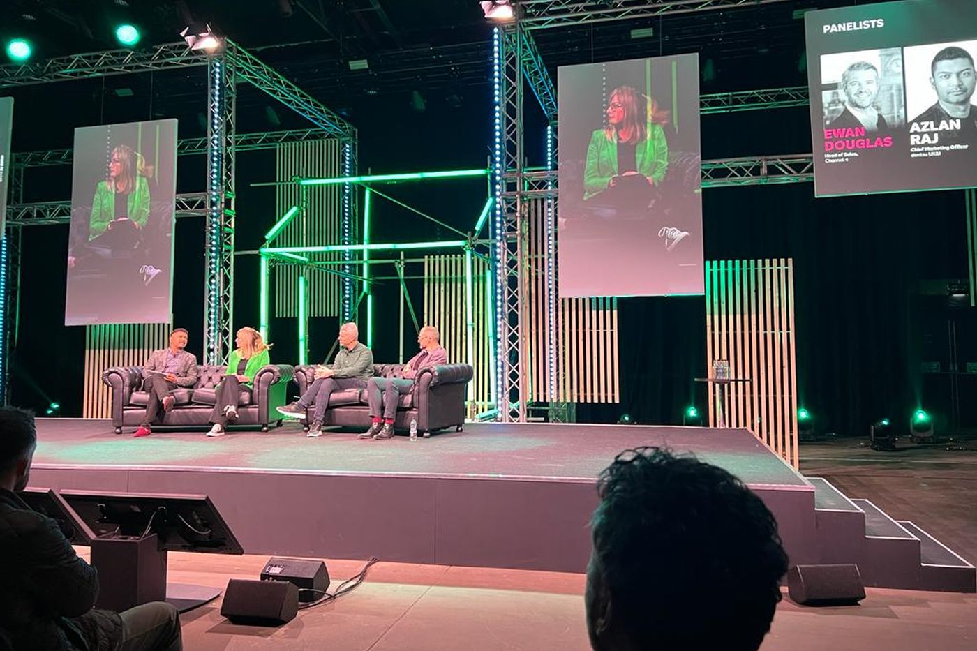 A four-person panel on sofas under green stage lighting, with the panellists named on the screen to the right.