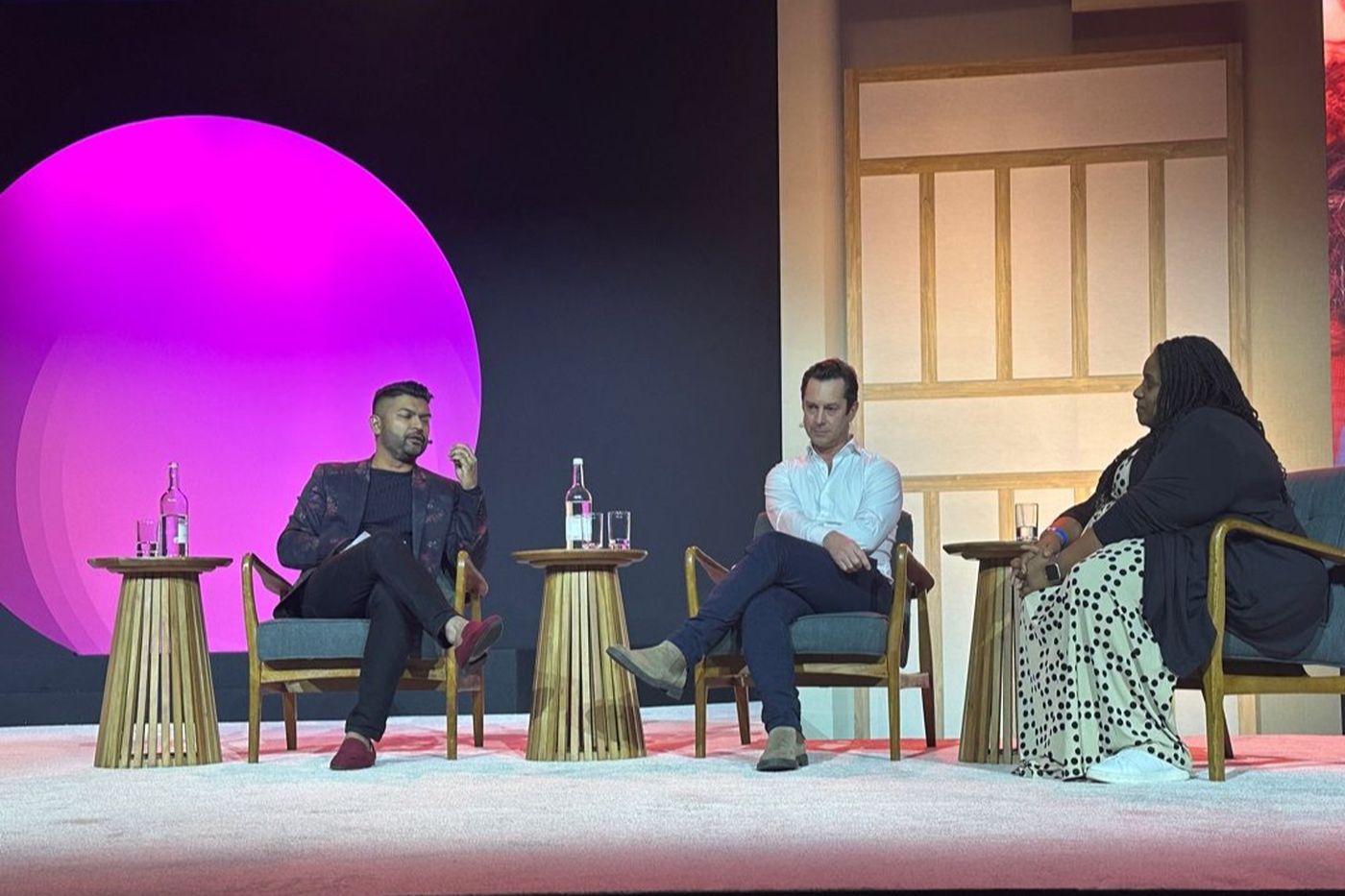 A three-person panel seated in armchairs on a pale stage, in front of a large magenta circle.