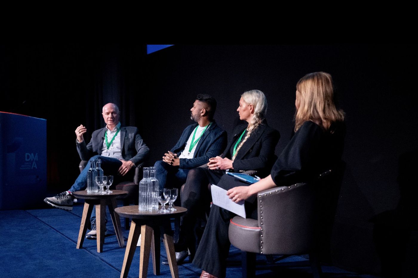 A four-person panel seated in conversation on a dark stage with a DMA backdrop.
