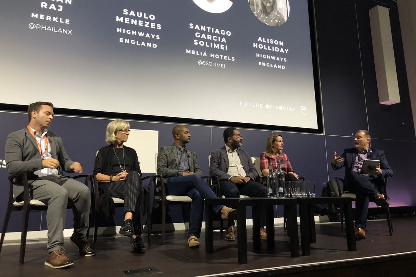 A six-person panel on stage, with the speakers' names and photographs on the screen behind them.
