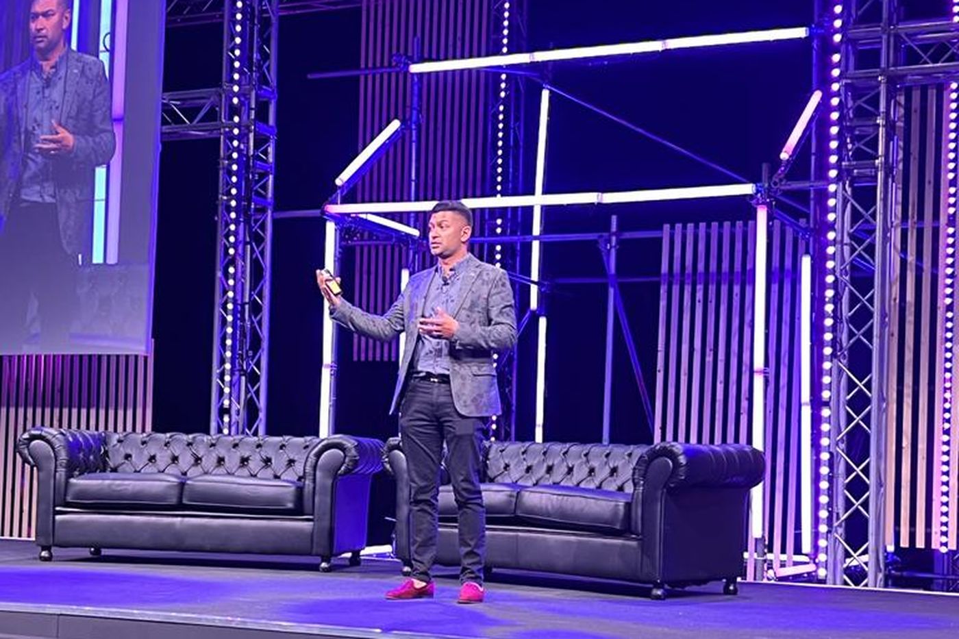 Azlan Raj speaking alone on a stage lit blue and purple, two leather sofas behind him.